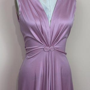 May include: A sleeveless, lilac dress with a deep V-neck. The waist is gathered and adorned with a circular embellishment. The fabric has a subtle sheen, and the dress appears to be made of a flowing material. The dress is a vintage style.
