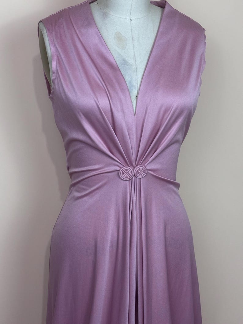 May include: A sleeveless, lilac dress with a deep V-neck. The waist is gathered and adorned with a circular embellishment. The fabric has a subtle sheen, and the dress appears to be made of a flowing material. The dress is a vintage style.