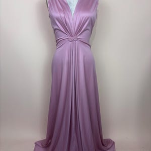 May include: A sleeveless, floor-length lavender dress. The dress has a deep V-neck and a gathered waist with two decorative buttons. The fabric drapes elegantly, creating a flowing silhouette. The dress is displayed on a mannequin.