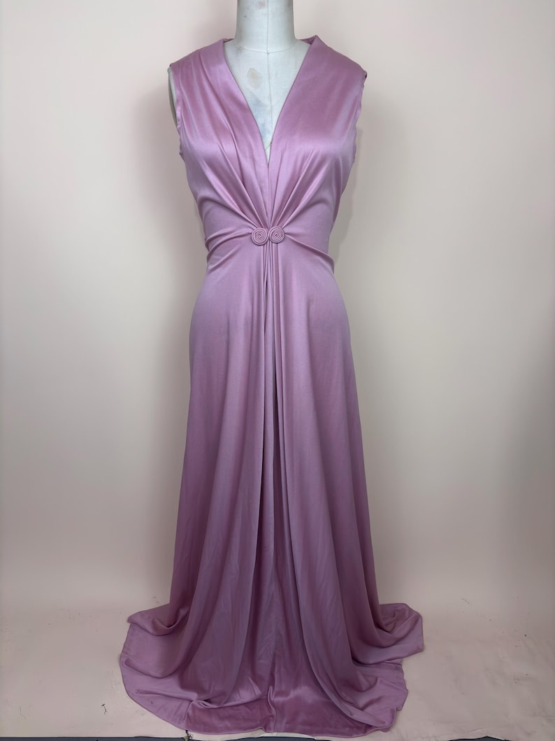 May include: A sleeveless, floor-length lavender dress. The dress has a deep V-neck and a gathered waist with two decorative buttons. The fabric drapes elegantly, creating a flowing silhouette. The dress is displayed on a mannequin.