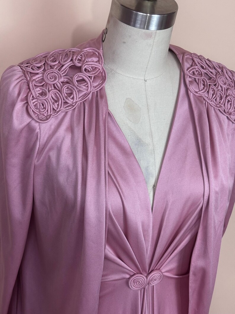 May include: A vintage, pale purple dress with long sleeves and a V-neck. The dress has decorative, circular embellishments on the shoulders and a matching circular clasp at the waist. The fabric has a satin-like sheen.