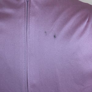 May include: A close-up shot of a lavender-coloured garment, possibly a dress or top, featuring a central zipper. The fabric has a smooth texture and a subtle sheen. Two small dark marks are visible on the fabric near the shoulder.