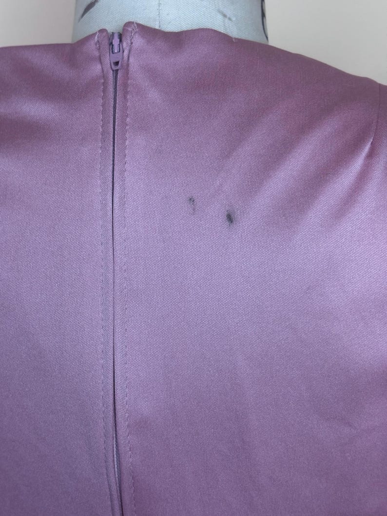 May include: A close-up shot of a lavender-coloured garment, possibly a dress or top, featuring a central zipper. The fabric has a smooth texture and a subtle sheen. Two small dark marks are visible on the fabric near the shoulder.