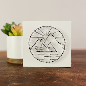 May include: A white square wooden plaque with a black and white string art design of a mountain range with a sun and trees. The design is in a circle.