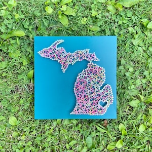 May include: A teal blue wooden board with a string art design of the state of Michigan. The string art is made with colorful threads in pink, yellow, blue, and white. A small heart is included in the design.