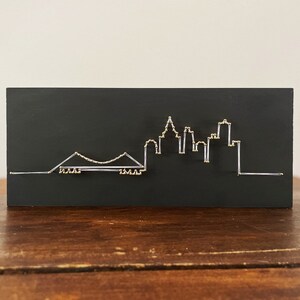 May include: A black wooden plaque with a white string art design of a city skyline with a bridge. The string art is outlined with gold beads.