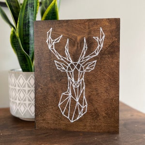 May include: A geometric deer head design created with white string wrapped around nails hammered into a dark brown wooden board.