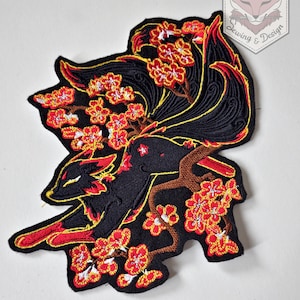 Op de afbeelding: Geborduurde patch met een zwarte vos met rode accenten, springend door een bloeiende kersenbloesemtak. De patch heeft een zwarte achtergrond met gele en rode details. De patch heeft de tekst "Karakitsunui Sewing & Design".