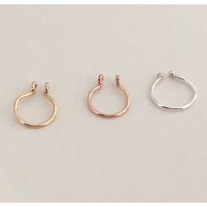 Puede incluir: Tres anillos de nariz en oro, oro rosa y plata. Cada anillo tiene un diseño simple y abierto con una pequeña bola o bucle en los extremos. Los anillos se muestran sobre un fondo claro.
