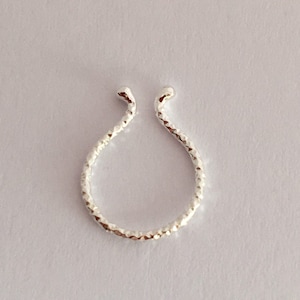 Puede incluir: Un anillo de nariz plateado con un diseño texturizado y martillado. El anillo de nariz tiene forma de U con dos puntas redondeadas y una banda texturizada. La joya está sobre un fondo blanco.
