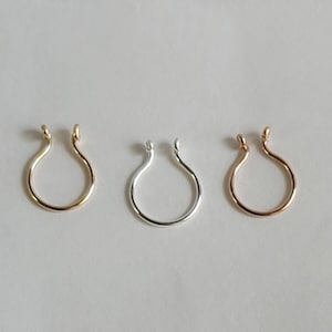 Puede incluir: Tres piercings de nariz en oro, plata y oro rosa. Cada anillo tiene un diseño en forma de U con extremos redondeados. Los anillos se muestran sobre una superficie blanca, mostrando sus diferentes acabados metálicos.