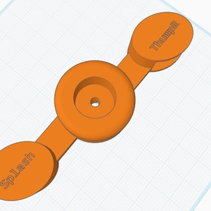 May include: Orange 3D-printed object with a central circular component and two oval-shaped end pieces. The end pieces are labeled "Splash" and "ThumpR". The object is set against a grid background.