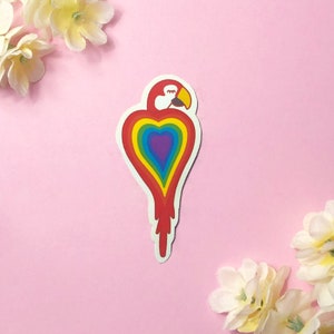 以下が含まれることがあります： 虹色のハート型のステッカーで、上部に赤いオウムの頭があります。オウムは左を向いていて、目は閉じられています。ハートは虹色で、中心は紫です。