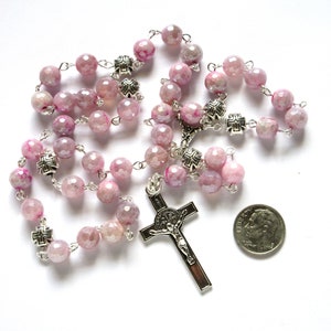May include: A rosary with faceted pink beads and silver-toned cross accents. The crucifix features a detailed depiction of Jesus. A dime is included for size reference. The beads are strung on a silver chain.