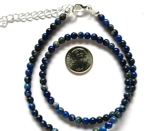 May include: A blue and grey gemstone beaded necklace with a silver clasp. The necklace is laid out on a white surface with a US dime for scale.