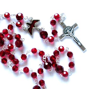 May include: A red crystal rosary with a silver crucifix and a silver dove charm. The crucifix has the words "AVE MARIA" on it.