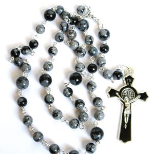 May include: A silver chain rosary with black and gray speckled beads and a silver crucifix with a black enamel background. The crucifix features a silver figure of Jesus Christ and the words "IN RI" on the top.