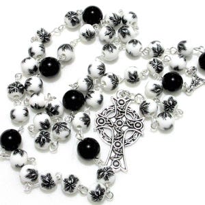 May include: A black and white rosary with a silver Celtic cross charm. The beads are white with a black snowflake design.