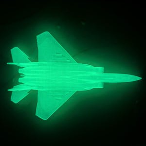 May include: A glowing green model airplane against a black background. The aircraft has a detailed design with wings, a fuselage, and tail fins. The model is illuminated, creating a striking contrast against the dark backdrop.