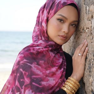 May include: A woman wearing a pink and purple tie-dye hijab and a black top. She is wearing gold bracelets and a ring. The background shows a stone wall and the ocean.