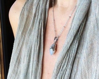 May include: A silver necklace with a pendant featuring a light blue crystal and a rectangular, clear stone. The necklace is worn over a light gray scarf.