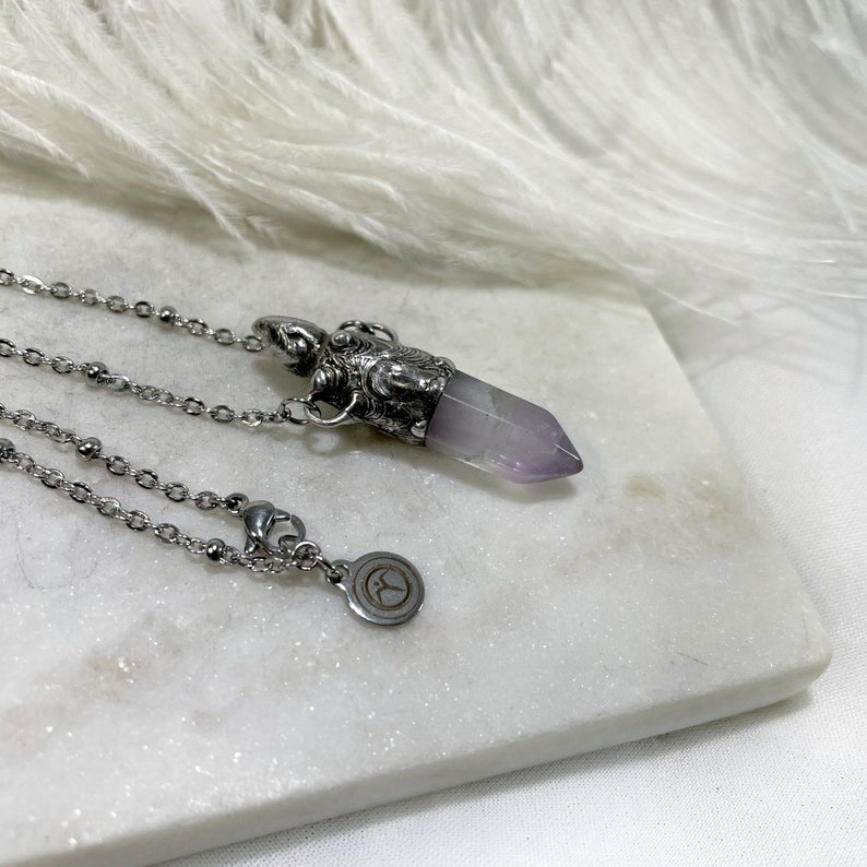 A silver chain necklace with a silver urn pendant featuring a purple amethyst crystal point. The pendant has an ornate design with a small silver charm attached to the chain.