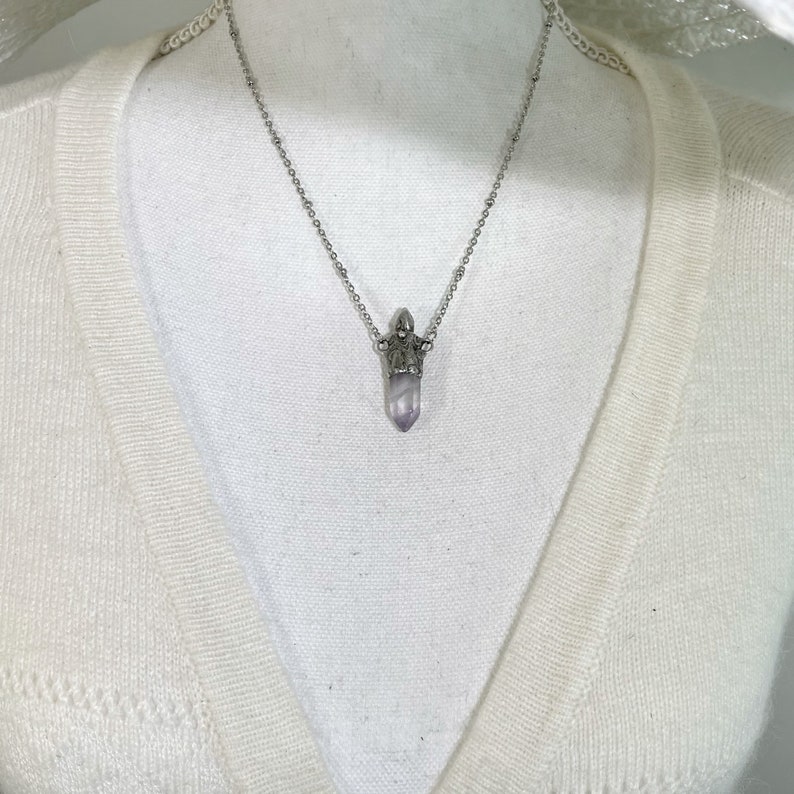 A silver chain necklace with a purple crystal urn pendant. The pendant is shaped like a point and is set in a silver crown-like setting.
