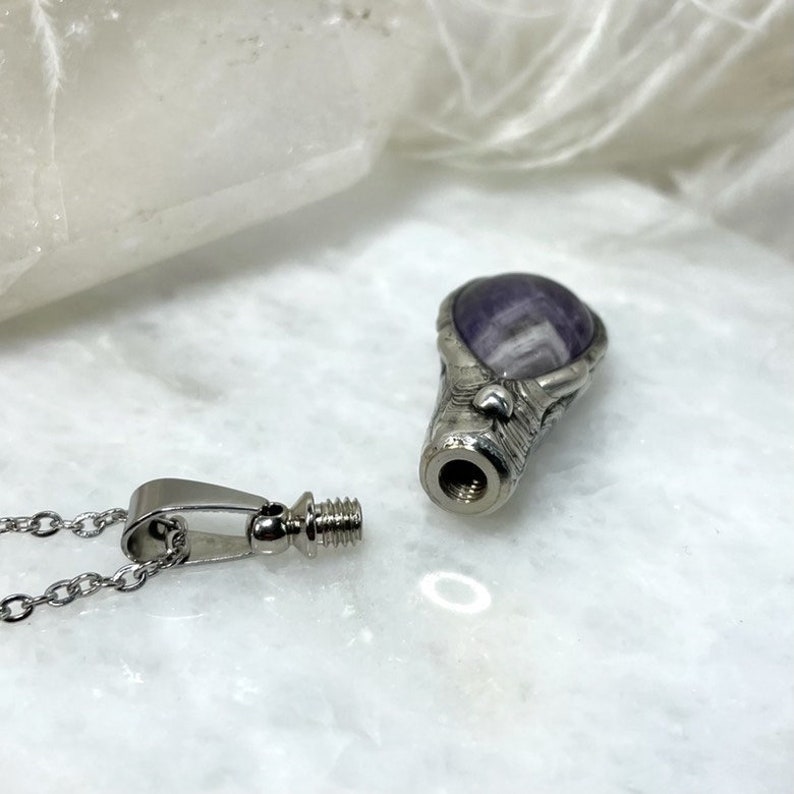 A silver pendant necklace with a purple amethyst stone. The pendant is shaped like a teardrop and has a detailed design. The top of the pendant is unscrewed showing that it has a container, suggesting it is memorial jewelry. The necklace is attached to a silver chain.