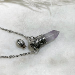 A silver chain necklace with a silver pendant that has a purple amethyst crystal on top. The pendant is shaped like a bottle with a screw-on top. The top is unscrewed and lying next to the pendant.