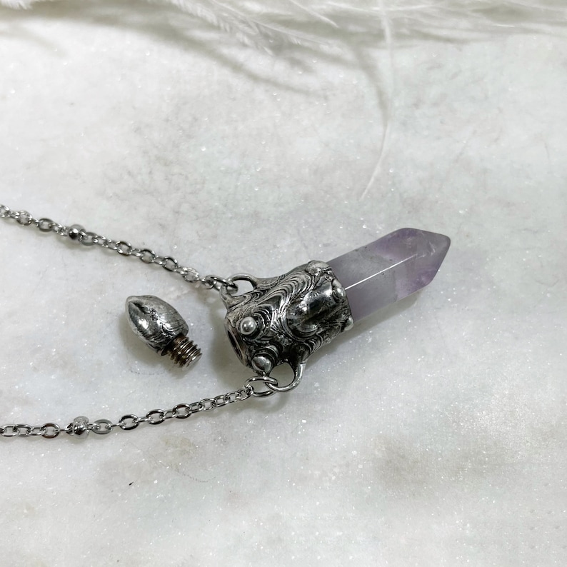 A silver chain necklace with a silver pendant that has a purple amethyst crystal on top. The pendant is shaped like a bottle with a screw-on top. The top is unscrewed and lying next to the pendant.