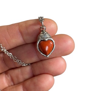 Può includere: Collana con pendente color argento con una pietra a forma di cuore, marrone-rossastro. La pietra è incastonata in una cornice d'argento decorativa, sospesa a una catena sottile. Il pendente ha un design ornato, con un motivo floreale o a foglie dettagliato.
