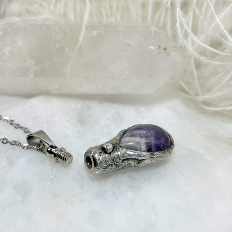 A silver urn pendant necklace with a purple amethyst stone set in a detailed silver design. The urn pendant is open on one end and has a screw top closure. The necklace is on a white surface.