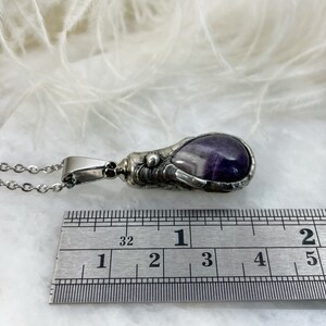 A silver pendant with a purple amethyst stone. The pendant is shaped like a claw and is hanging from a silver chain. The pendant is measured against a ruler showing its size which is 1.5”
