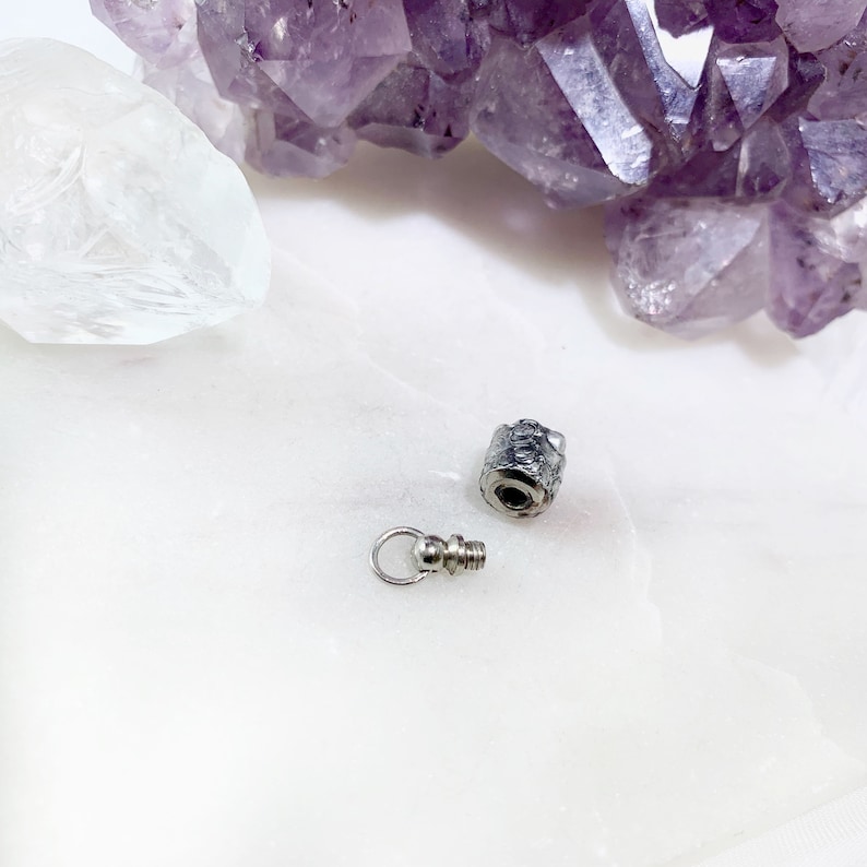 May include: A silver bottle stopper with a threaded top and a small loop for attaching to a chain. The stopper is on a white surface.