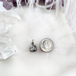 May include: A silver charm in the shape of a small, textured bell. The charm is about the size of a US dime.