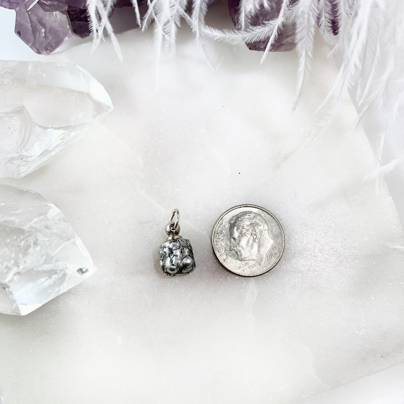 May include: A silver charm in the shape of a small, textured bell. The charm is about the size of a US dime.