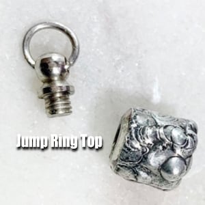 May include: Silver bottle stopper with a jump ring top and a detailed, textured design. The top of the stopper has a threaded screw.