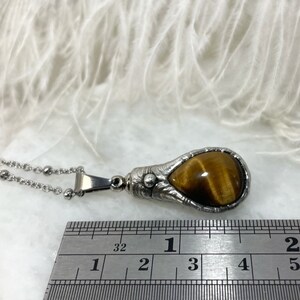 May include: A silver pendant necklace with a teardrop-shaped tiger's eye stone set in a detailed silver frame. The pendant measures approximately 1 inch in length. The necklace is made of a silver chain with small round beads.