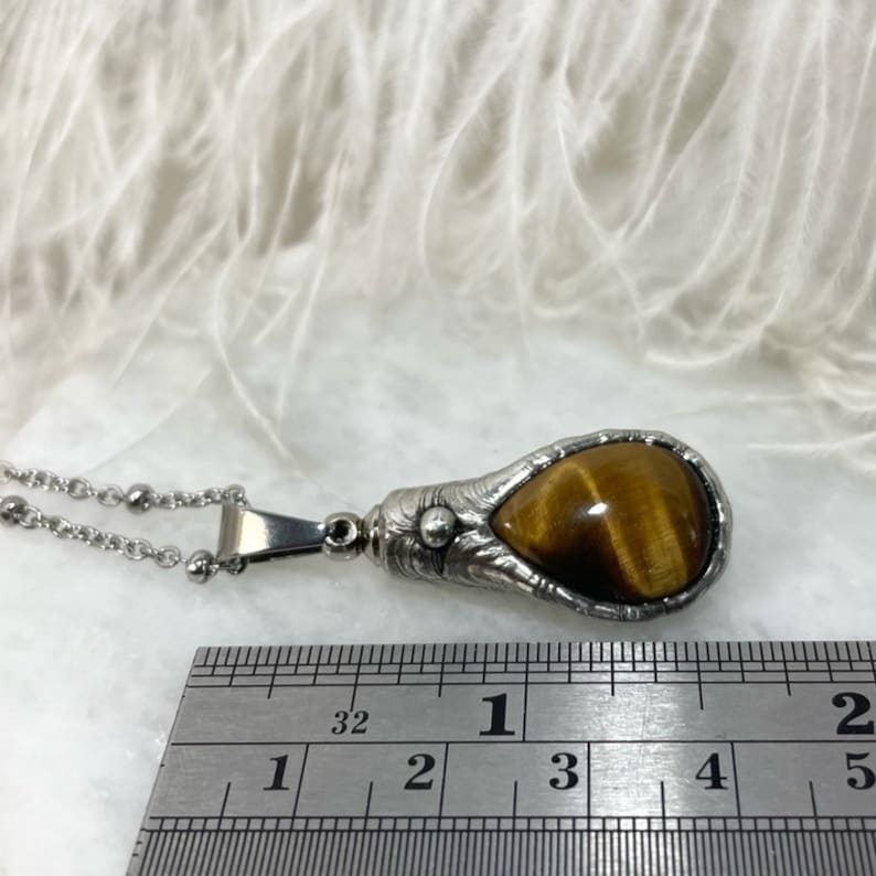 May include: A silver pendant necklace with a teardrop-shaped tiger's eye stone set in a detailed silver frame. The pendant measures approximately 1 inch in length. The necklace is made of a silver chain with small round beads.