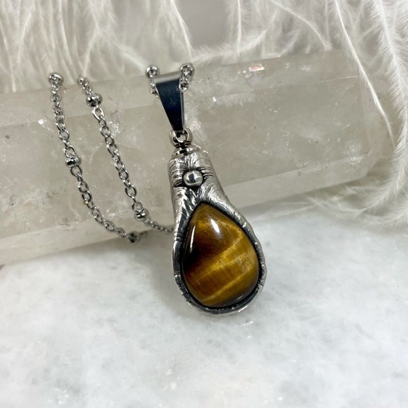 May include: A silver chain necklace with a teardrop-shaped pendant featuring a brown tiger's eye stone set in a silver bezel.