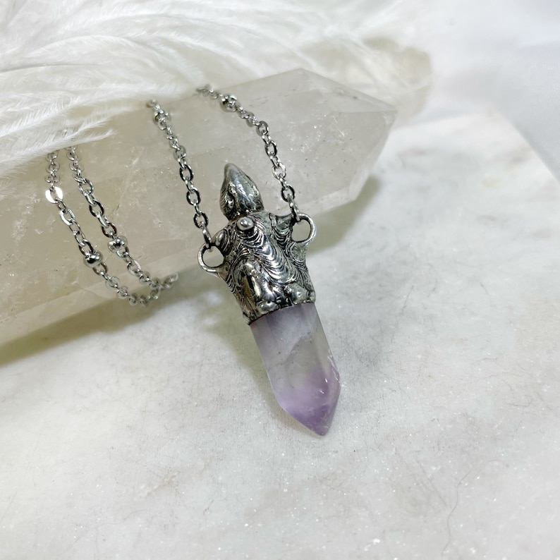 A silver necklace with a silver cremation urn pendant featuring a detailed design. The pendant is attached to a purple amethyst crystal point.