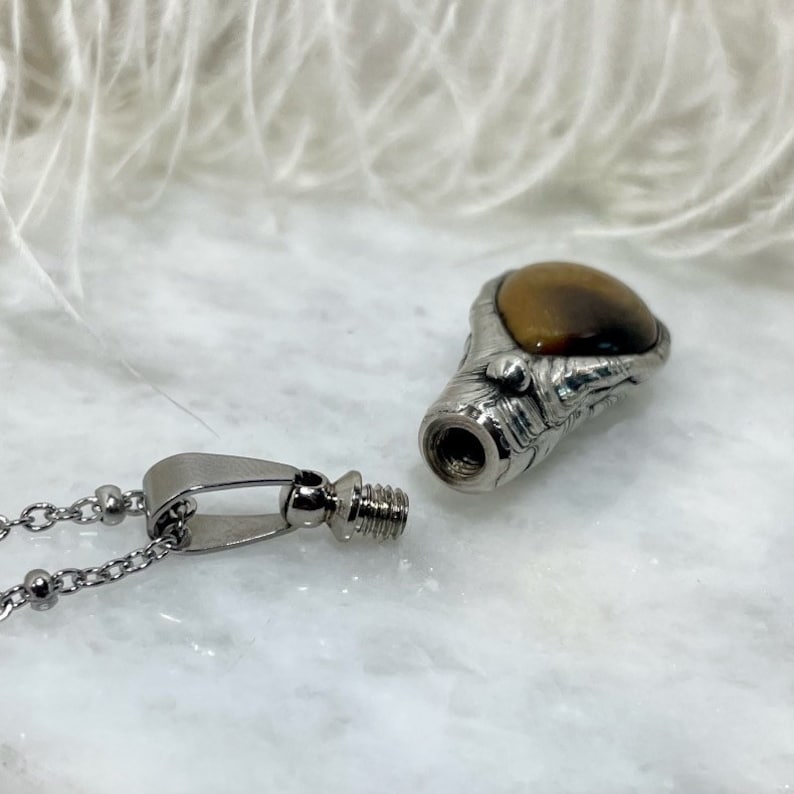 May include: A silver pendant necklace with a tiger's eye stone. The pendant is shaped like a claw and has a threaded opening. The necklace is made of a silver chain with small round beads.
