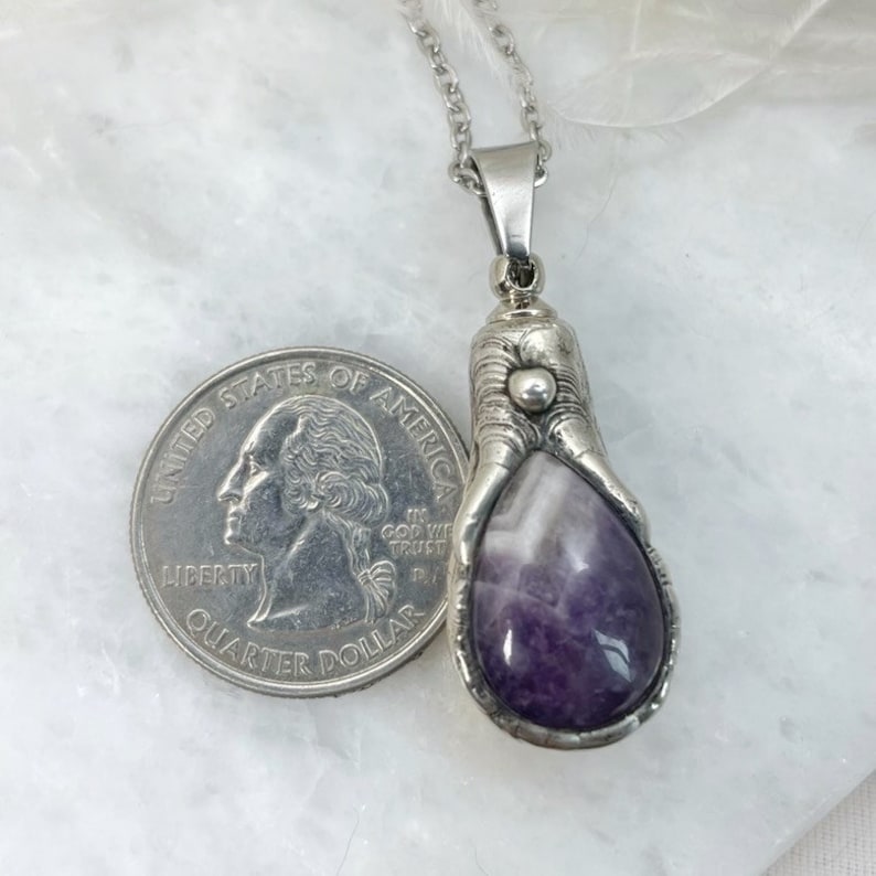 A silver pendant with a purple amethyst stone set in a claw design. The pendant is hanging from a silver chain and is next to a US quarter for size comparison.