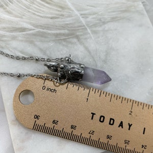 A silver cremation pendant necklace with a purple amethyst crystal point. The pendant is shaped like a small, ornate box with a silver chain attached. The necklace is lying next to a ruler. The ruler is marked in inches and centimeters and shows that the entire pendant is close to 1.5”.