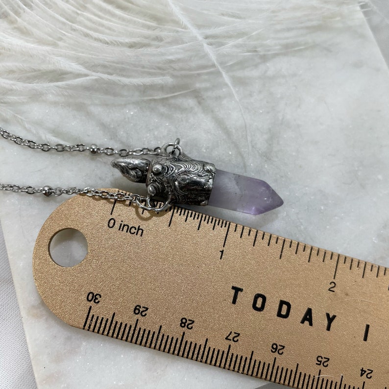 A silver cremation pendant necklace with a purple amethyst crystal point. The pendant is shaped like a small, ornate box with a silver chain attached. The necklace is lying next to a ruler. The ruler is marked in inches and centimeters and shows that the entire pendant is close to 1.5”.