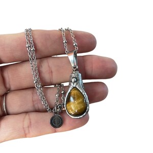 May include: A silver chain necklace with a teardrop-shaped pendant featuring a brown and gold tiger's eye stone.
