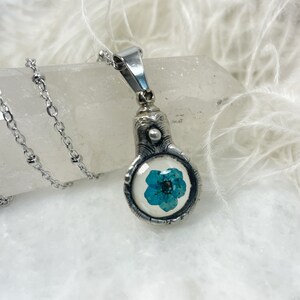Op de afbeelding: Een zilveren ketting met een hanger met een blauwe bloem geperst in een glazen koepel.
