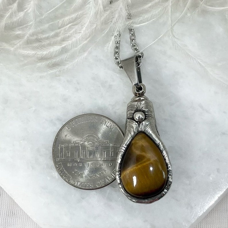 May include: A silver pendant necklace with a teardrop-shaped tiger's eye stone set in a silver bezel. The pendant is about the size of a US quarter.