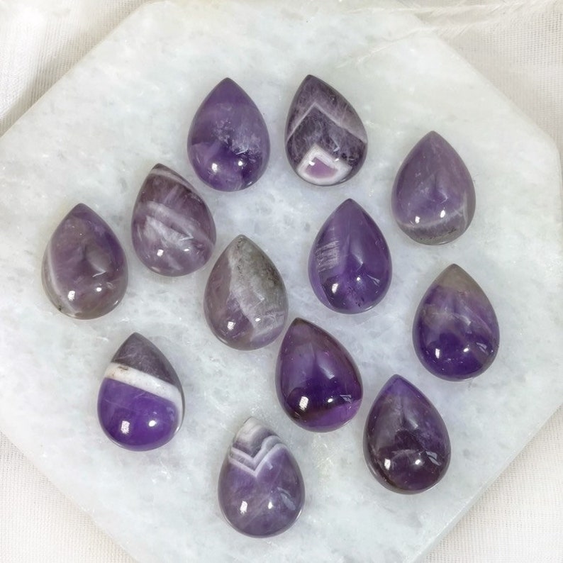 Twelve purple amethyst teardrop cabochons with white and gray inclusions. The cabochons are arranged in a semi-circle on a white surface.