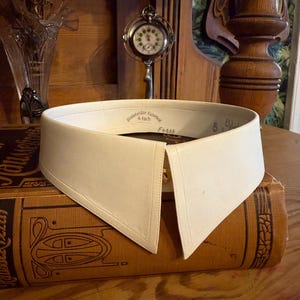 Puede incluir: Un cuello desmontable blanco roto con una parte delantera puntiaguda, apoyado en un libro antiguo marrón. El cuello tiene un cierre de botón y una etiqueta con texto. Un pequeño reloj y un jarrón con flores secas están en el fondo.